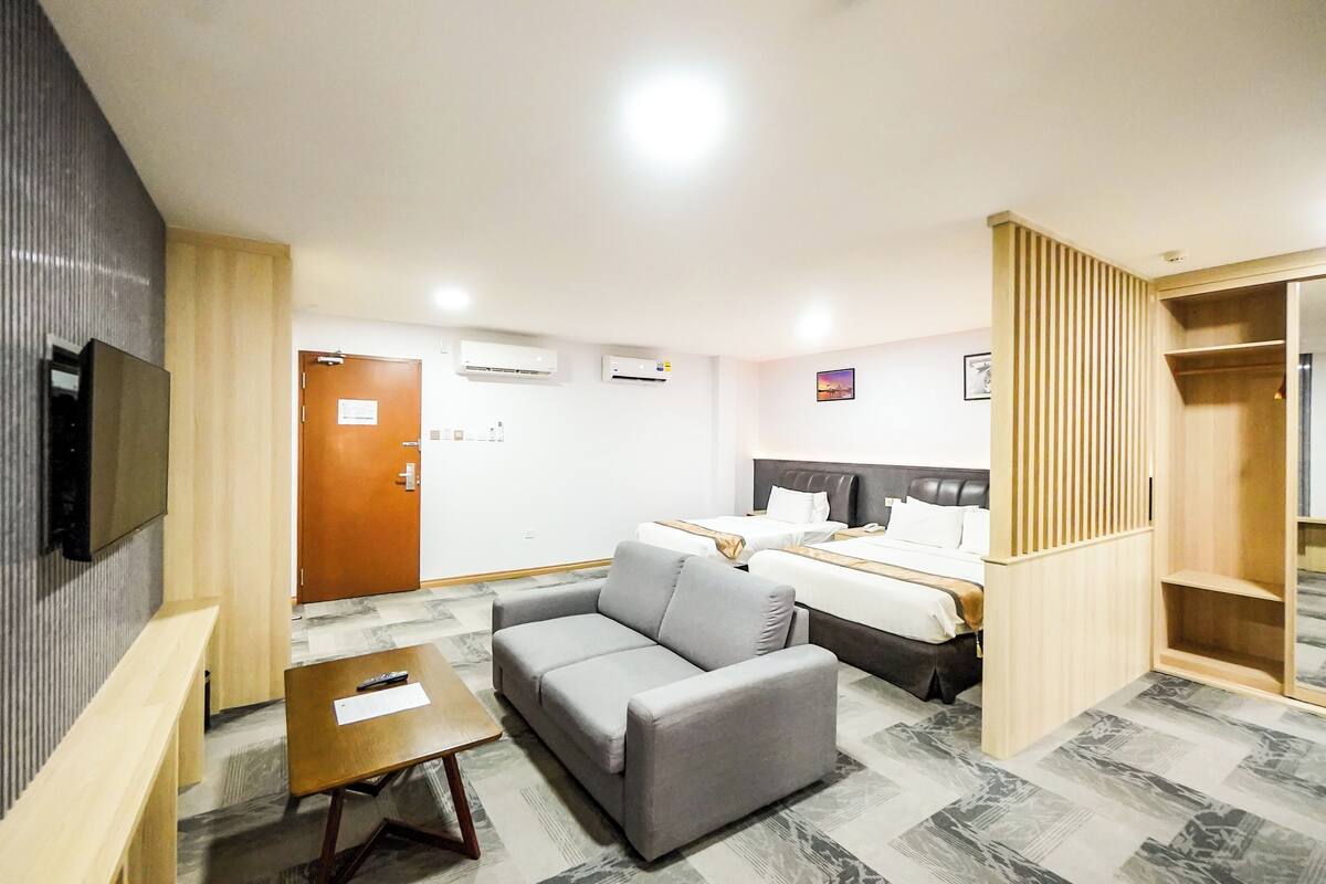 A hotel room with a grey sofa, a wooden table, a bed with white bedding, and a wardrobe.