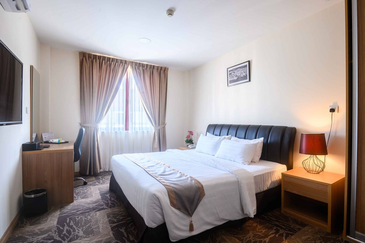 A hotel room with a large bed, a desk, a chair, a nightstand with a lamp, and a window with curtains.