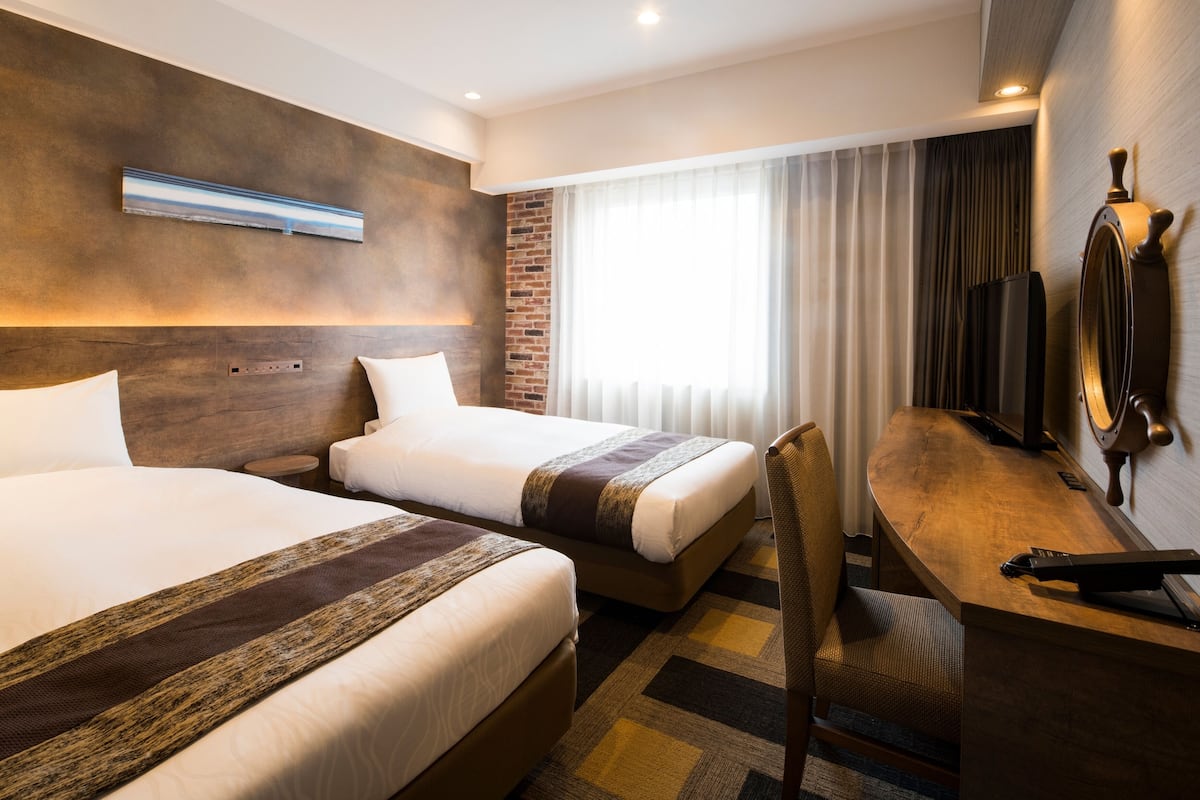 Ein Hotelzimmer mit zwei Betten, einem Schreibtisch, einem Stuhl, einem Fernseher und einem fenster mit Vorhängen.
