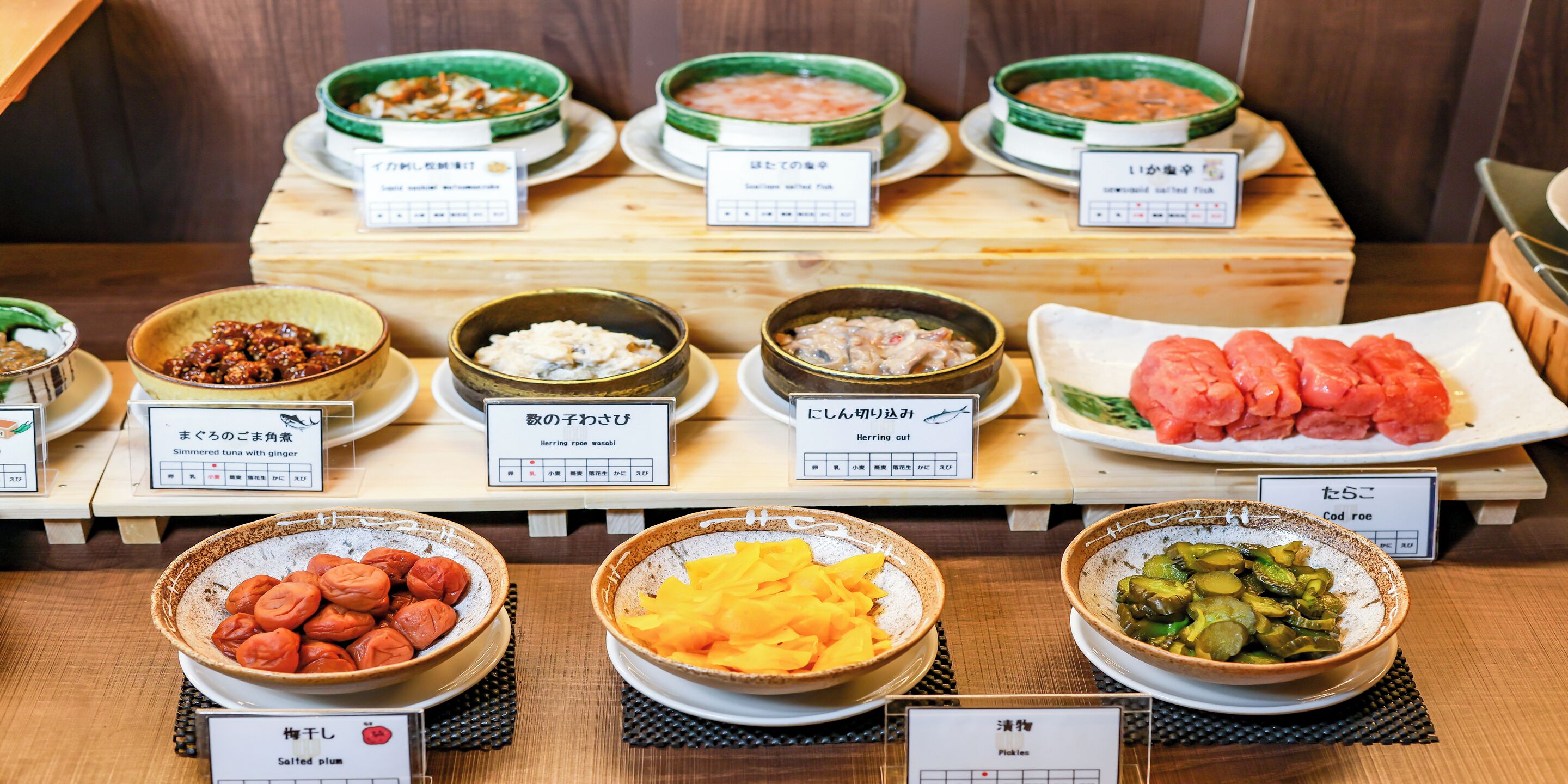 A variety of Japanese dishes displayed on a wooden table with labels in Japanese.