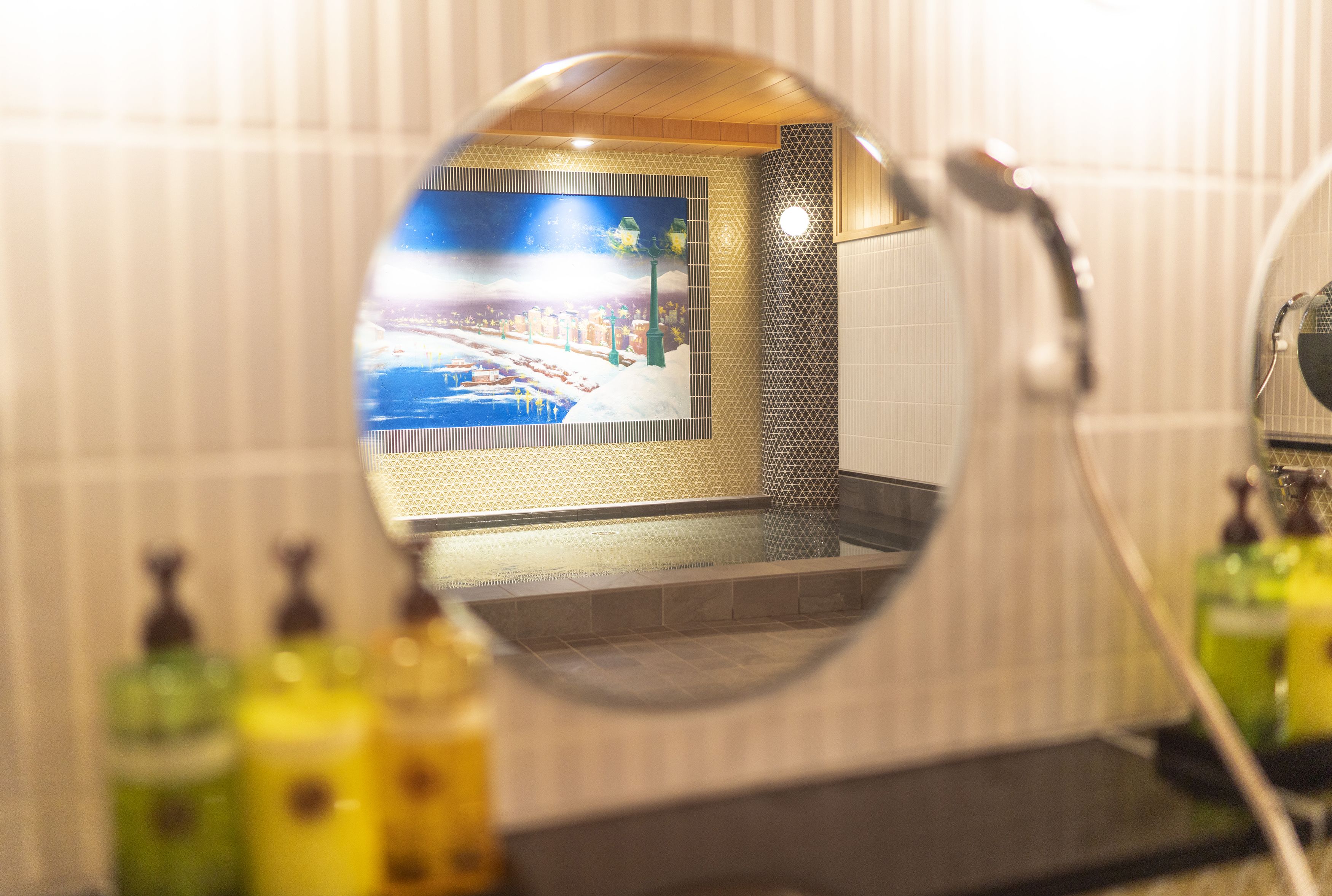 A bathroom with a round mirror reflecting a tiled wall and a window with a view of a cityscape.