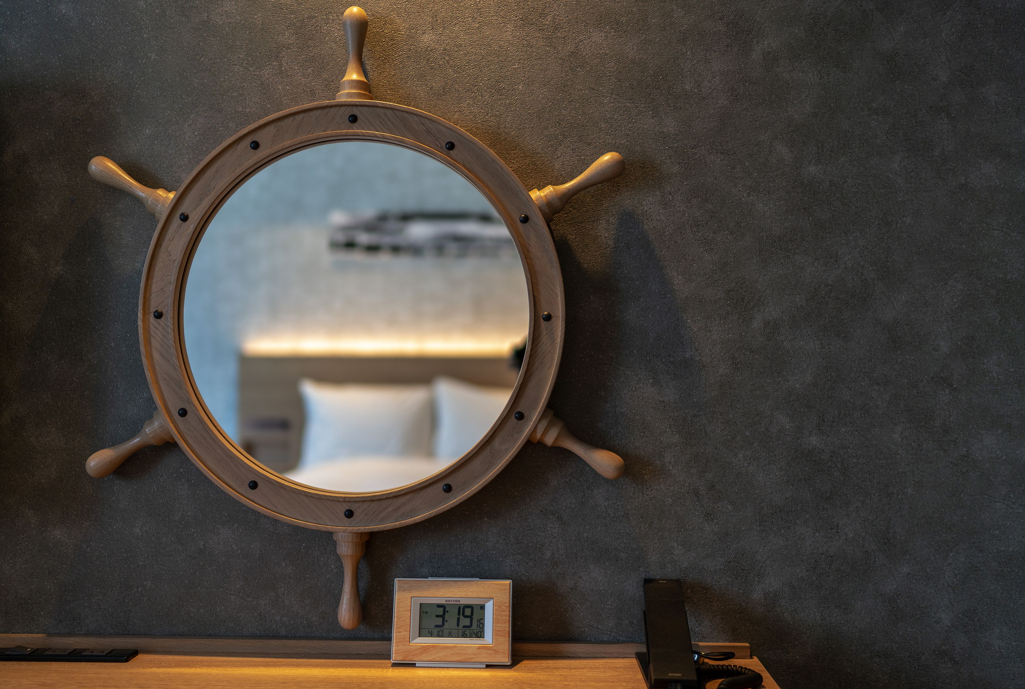 A round mirror with a ship's wheel design, mounted on a textured wall. Below the mirror, a digital clock displays the time.