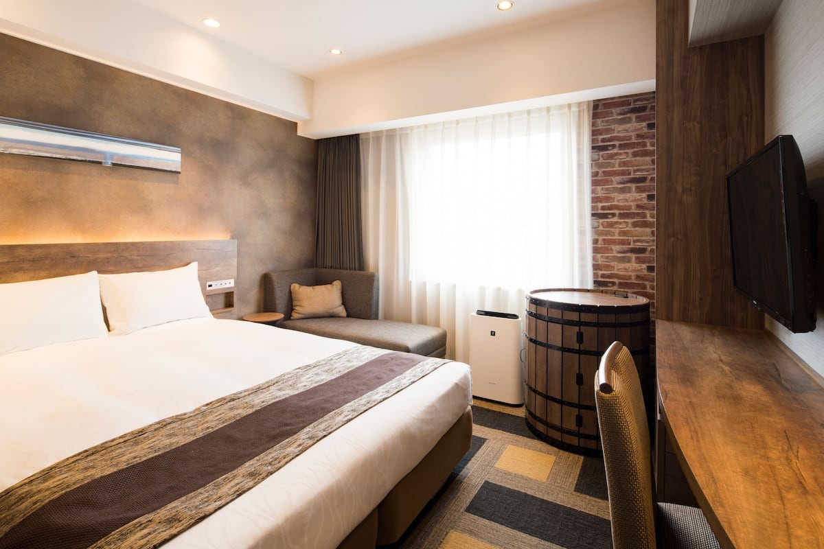 Ein Hotelzimmer mit einem großen Bett, einem hölzernen Kopfteil, einem kleinen Schreibtisch, einem Sessel, einem Fernseher, einem Fenster mit Vorhängen und einer Ziegelwand.
