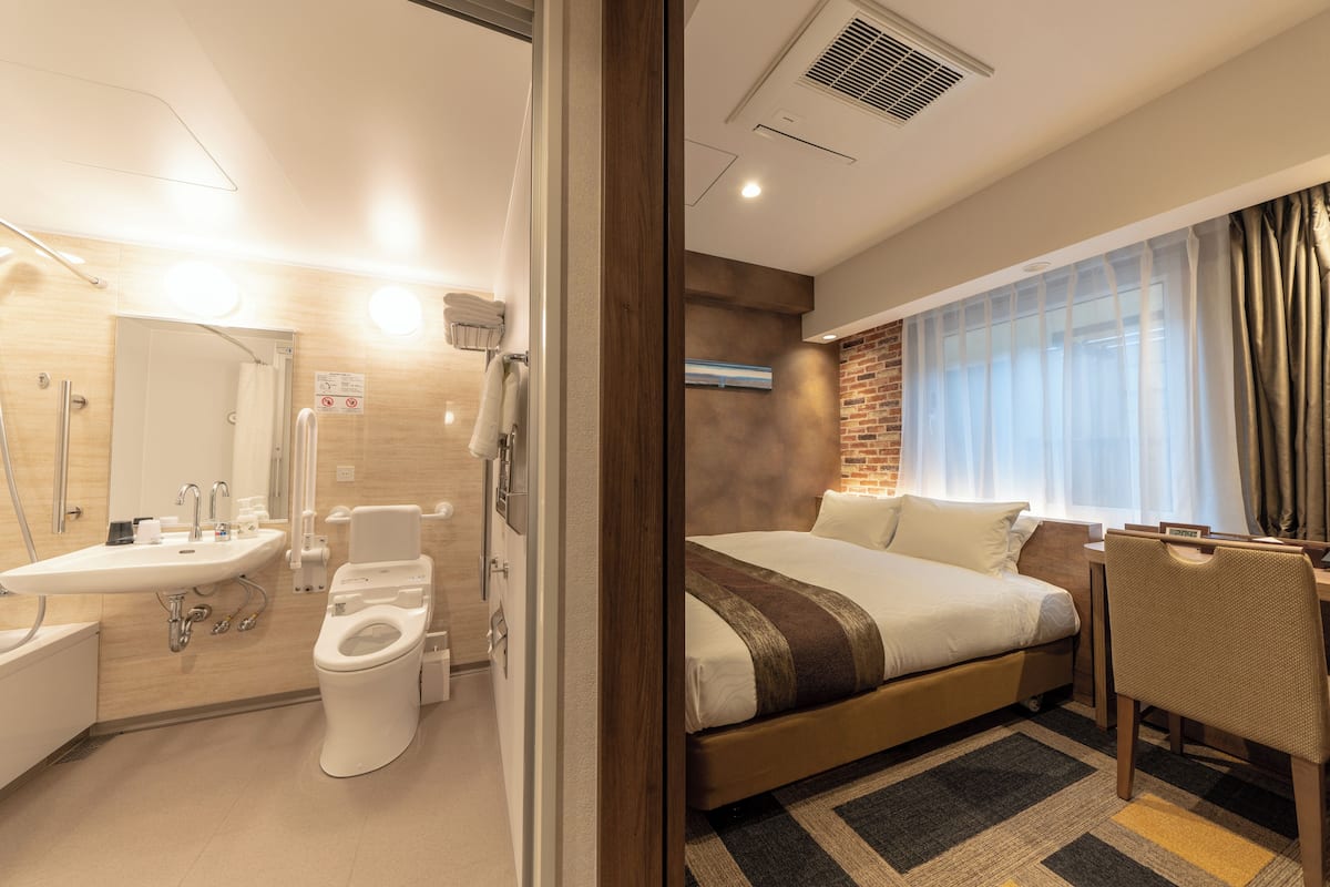 Ein modernes Hotelzimmer mit einem großen Bett, einem Schreibtisch, einem Sessel, einem Badezimmer mit Waschbecken und Toilette sowie einem Fenster mit Vorhängen.