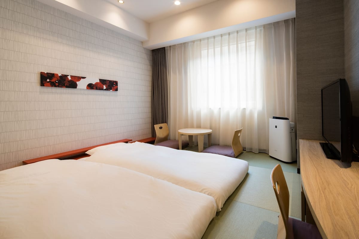 Ein Hotelzimmer mit zwei Betten, einem kleinen Tisch, einem Stuhl, einem Fernseher und einem Fenster mit Vorhängen.