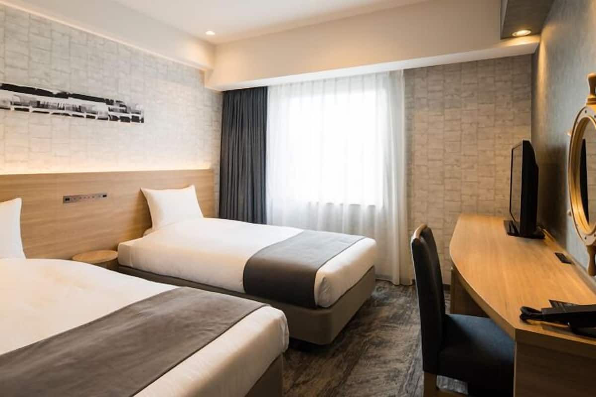 Ein Hotelzimmer mit zwei Betten, einem Schreibtisch, einem Stuhl, einem Fernseher und einem Fenster mit Vorhängen.