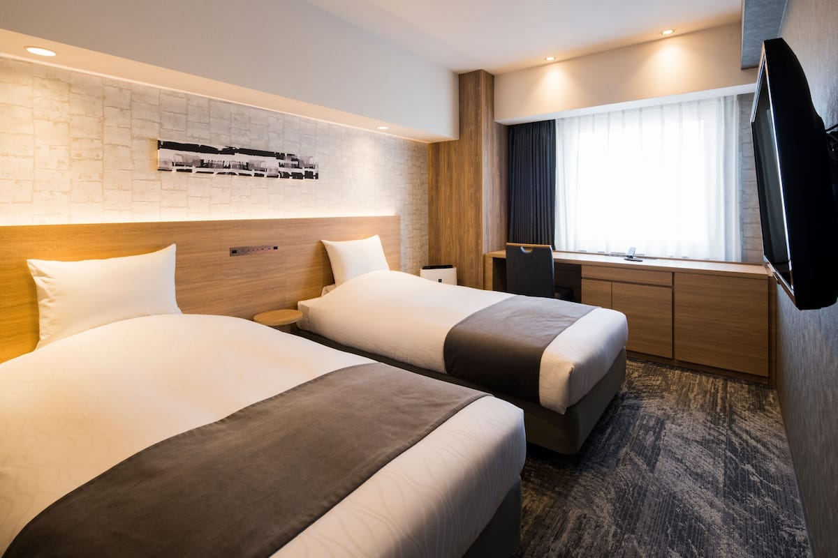 Ein Hotelzimmer mit zwei Betten, einem Fernseher, einem Fenster mit Vorhängen und einer Ziegelwand mit Fotoausstellung.