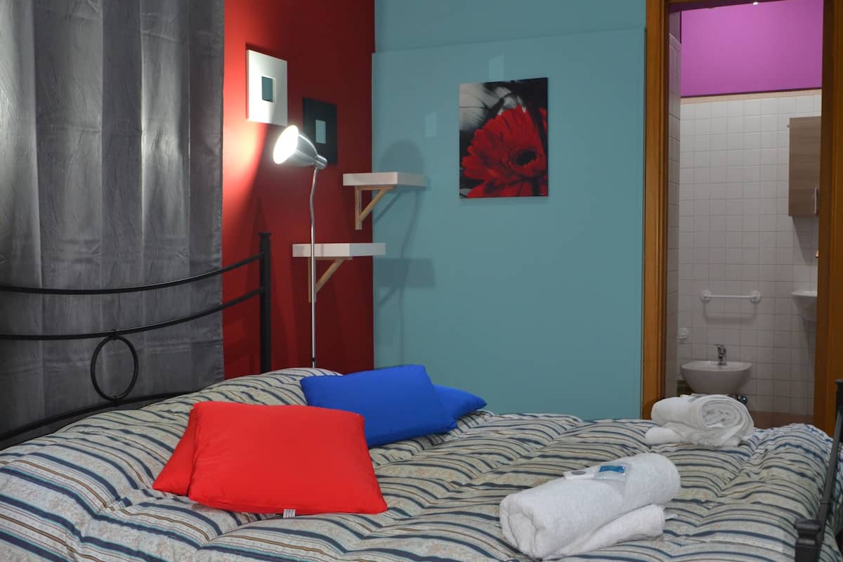 Una cama bien hecha con ropa de cama a rayas, un almohadón rojo y un almohadón azul. Hay una lámpara en una mesita de noche, un cuadro en la pared y un baño visible a través de una puerta abierta.