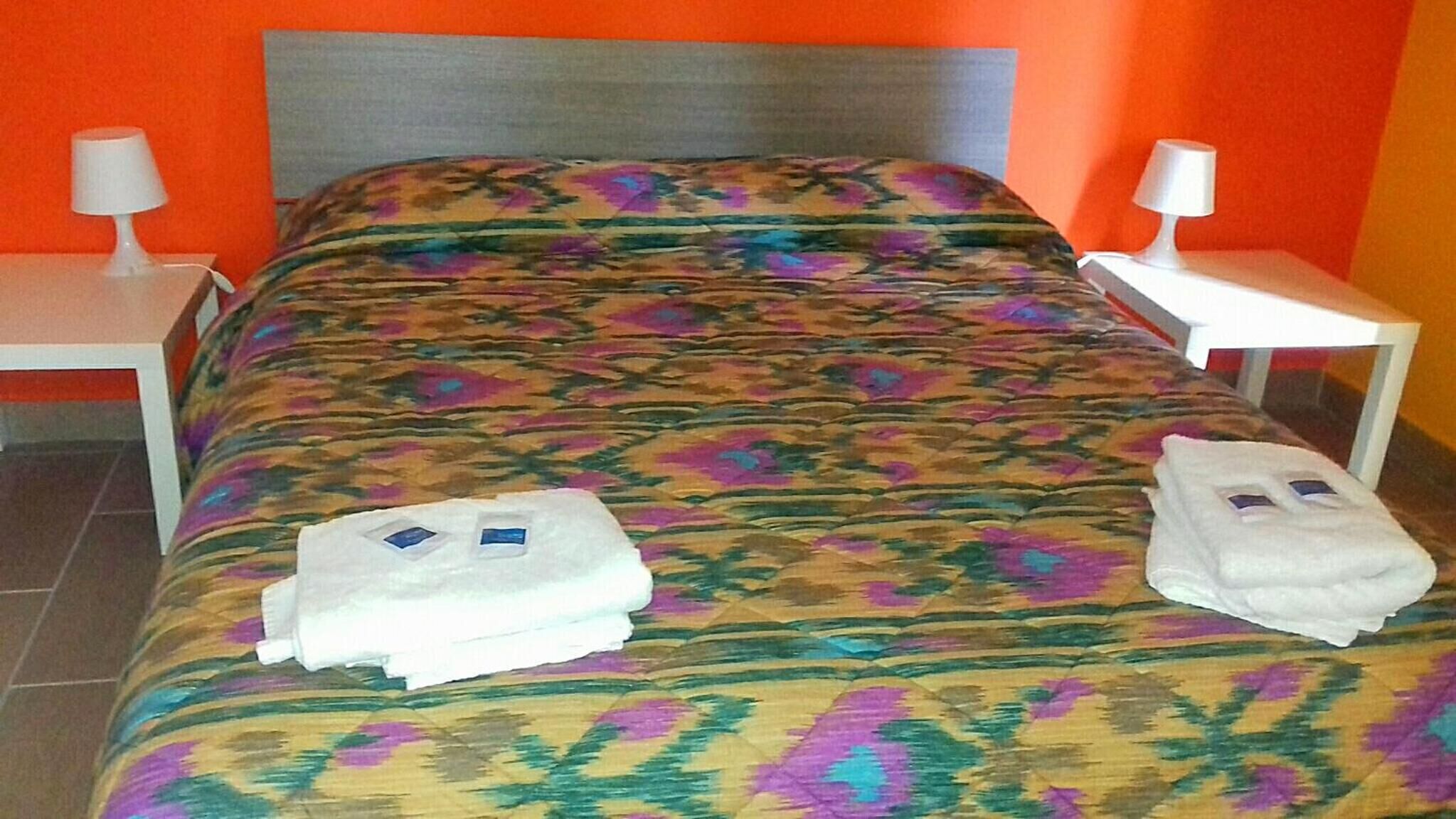 Una cama con colcha estampada, dos toallas con motivos faciales y dos lámparas de noche sobre mesitas blancas.
