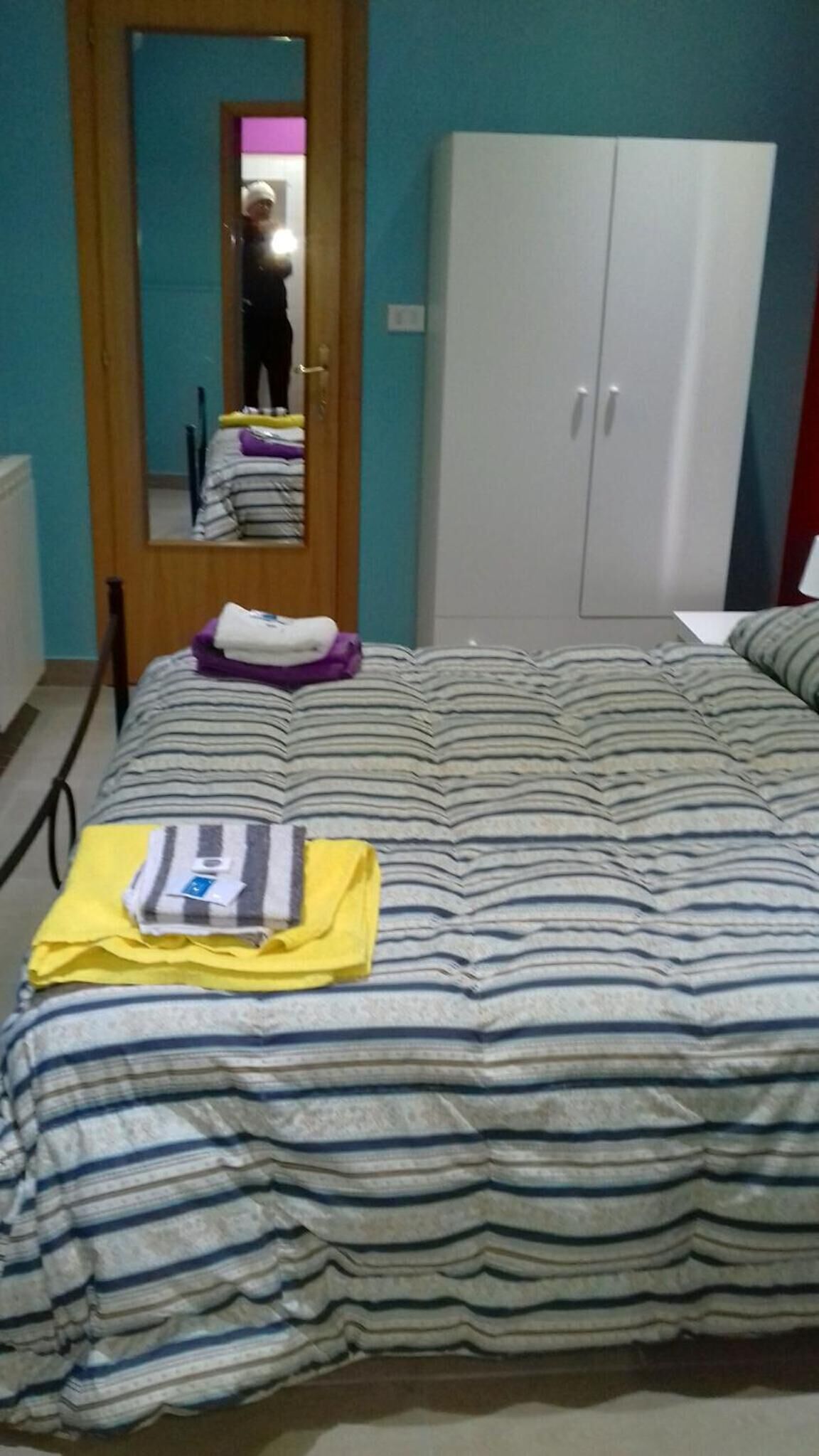 Quarto com colcha listrada, guarda-roupa e espelho refletindo uma pessoa.
