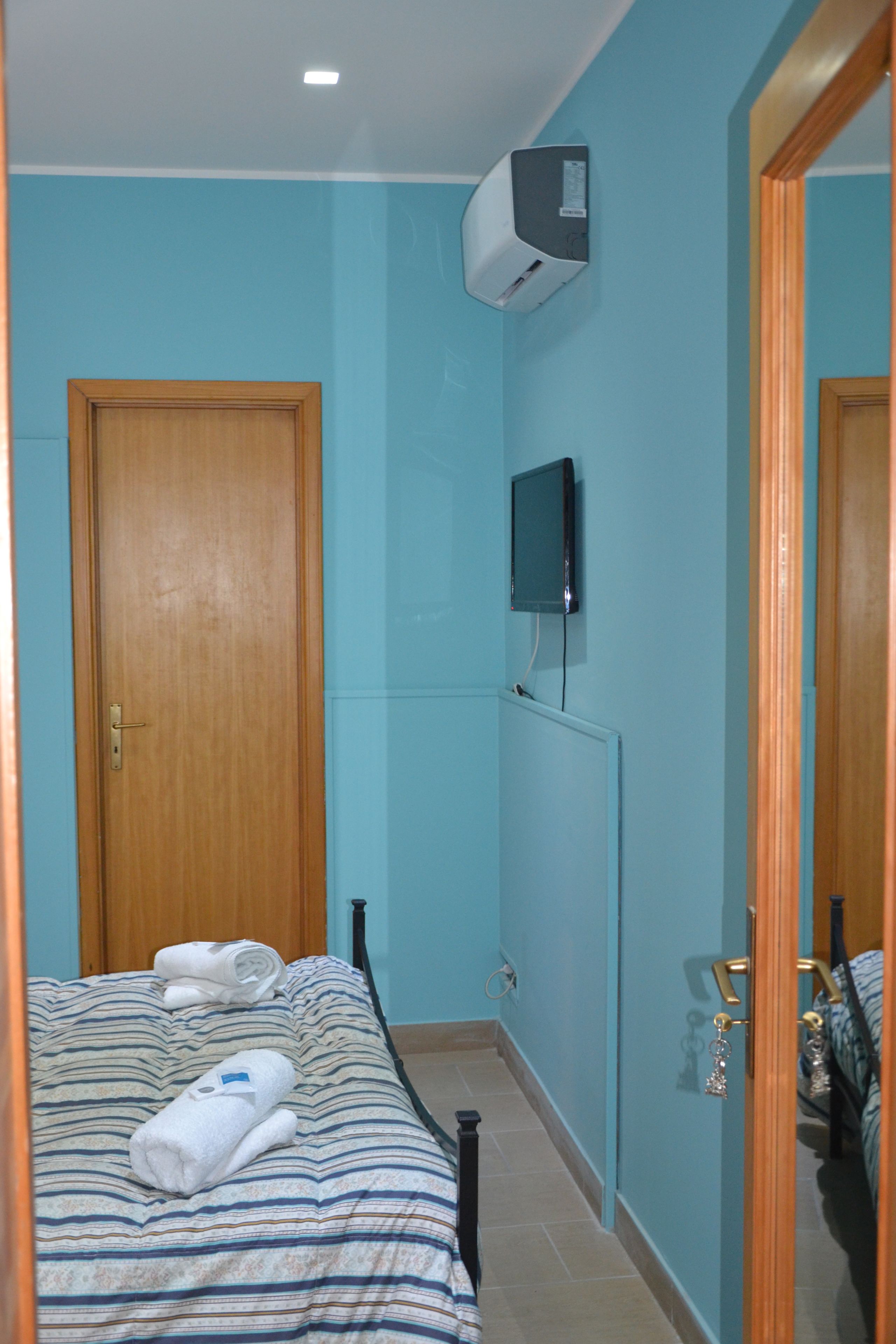 Una habitación de hotel pequeña y estrecha con una cama individual, una puerta de madera, un televisor montado y un equipo de aire acondicionado en la pared.