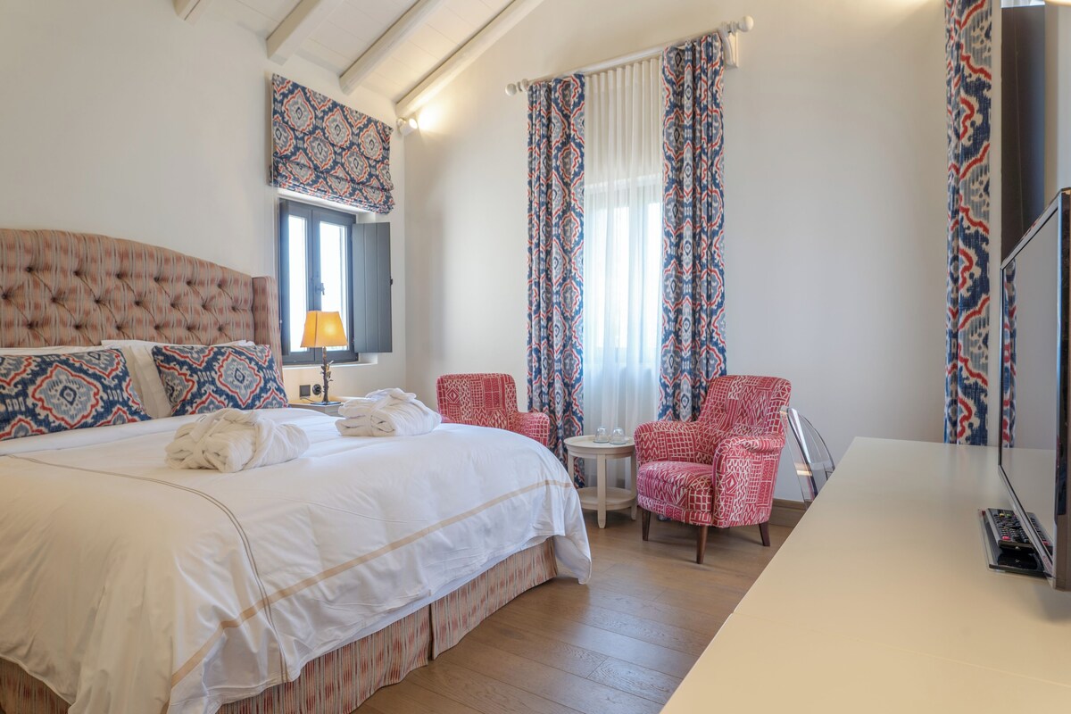 Habitación de hotel con una cama grande, una mesita redonda pequeña, un televisor y dos sillones estampados.