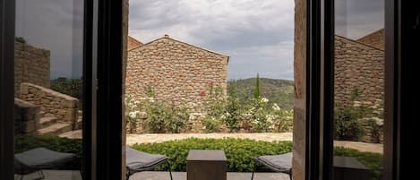 On aperçoit, à travers une porte vitrée, un bâtiment en pierre. Au premier plan, on distingue une table carrée et deux chaises.
