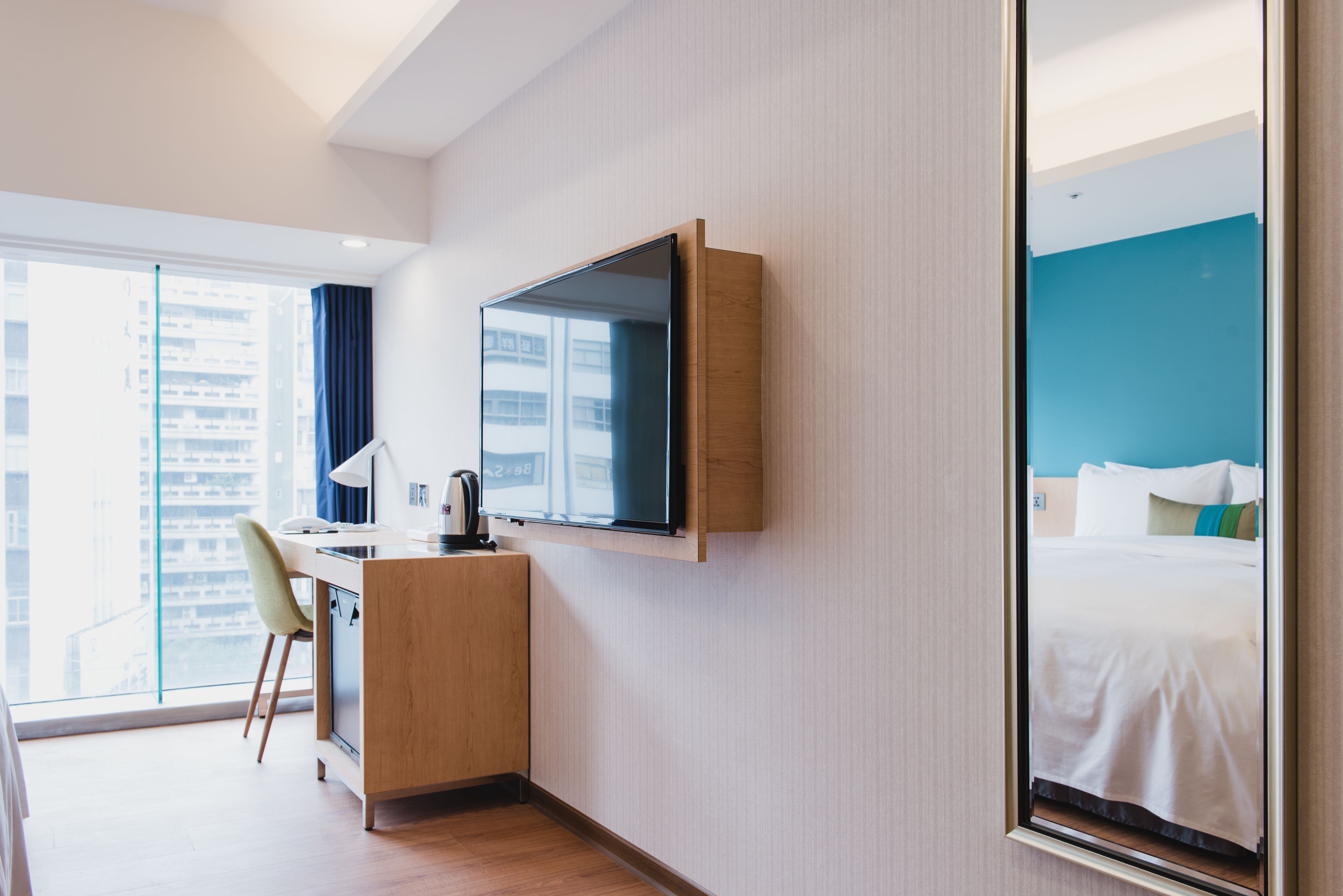 Quarto de hotel com cama, escrivaninha e cadeira, televisão na parede e uma grande janela com vista para a cidade.