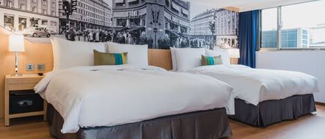 A hotel room with two beds, a large cityscape mural on the wall, a nightstand with a lamp, and a view of the city through the window.