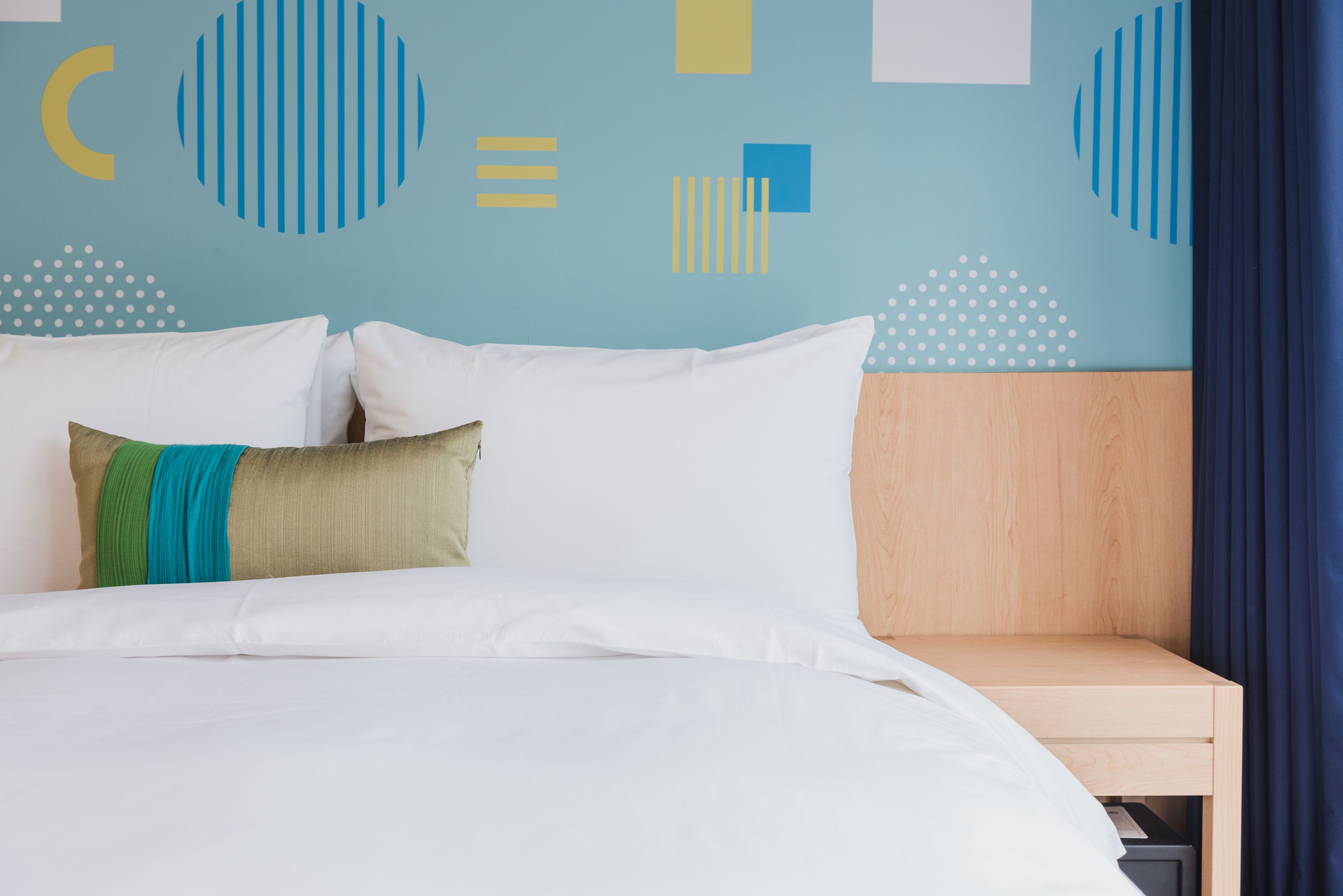 Cama bem arrumada com roupa de cama branca e almofada listrada em verde e azul, mesa de cabeceira de madeira e parede com padrões geométricos abstratos.