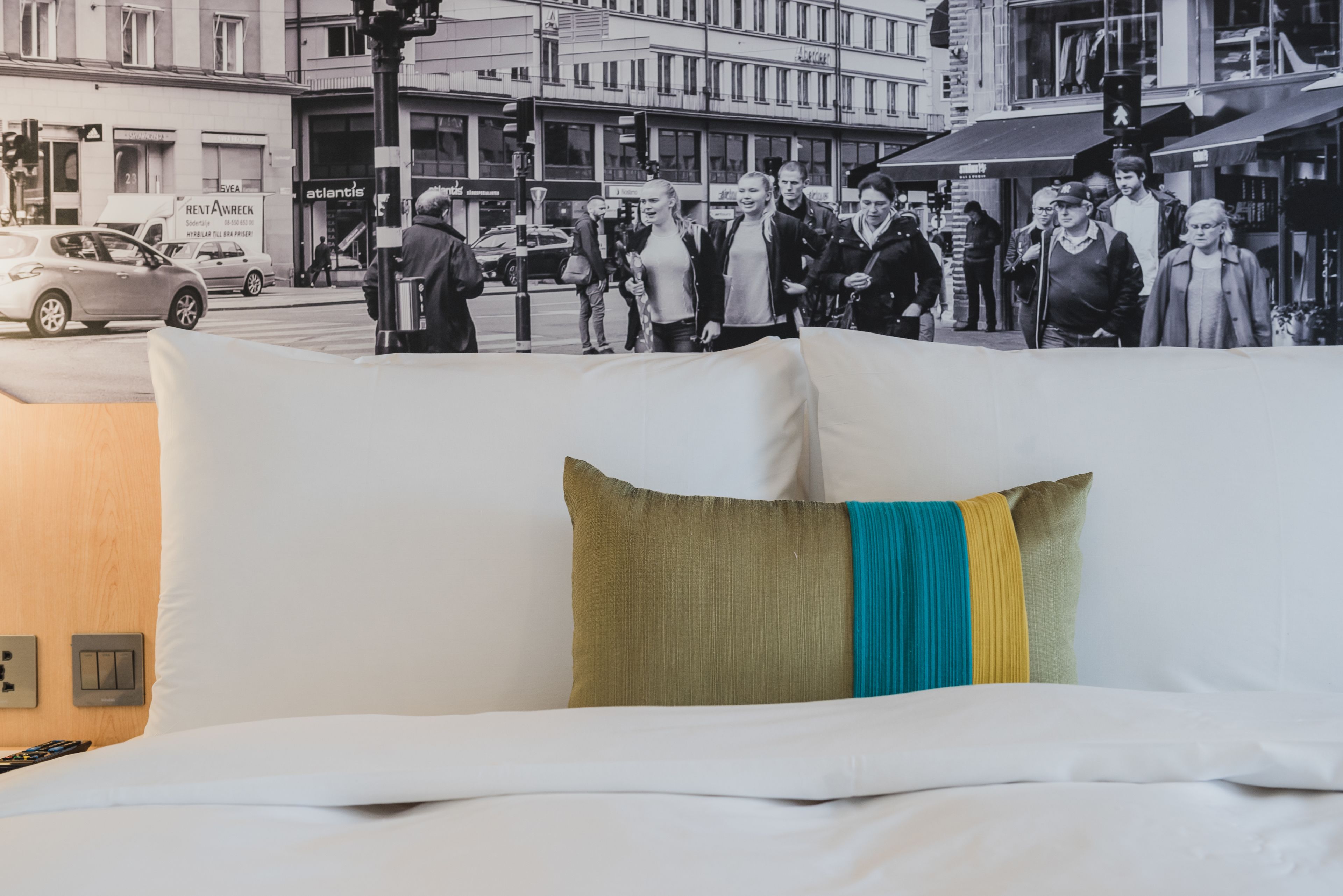 A hotel room with a large bed, a wall-mounted light switch, and a view of a busy street with pedestrians.