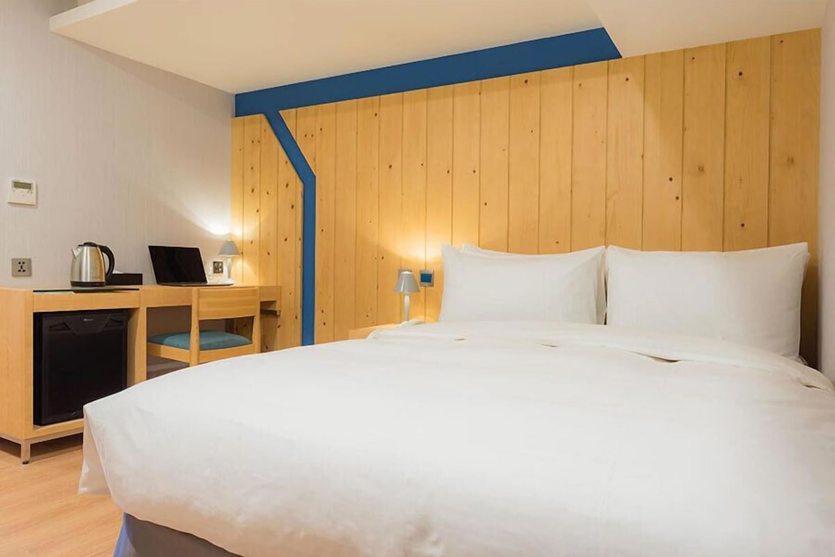 A hotel room with a large bed, a desk with a laptop, a kettle, and a small refrigerator.