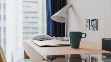 Habitación de hotel con un escritorio de madera, una laptop, un cuaderno, un bolígrafo, una taza verde azulada, una lámpara blanca y un teléfono.