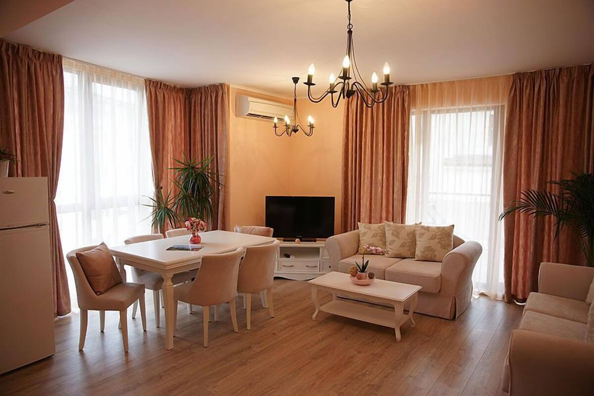 A modern living room with a dining area, a flat-screen TV, and a chandelier.