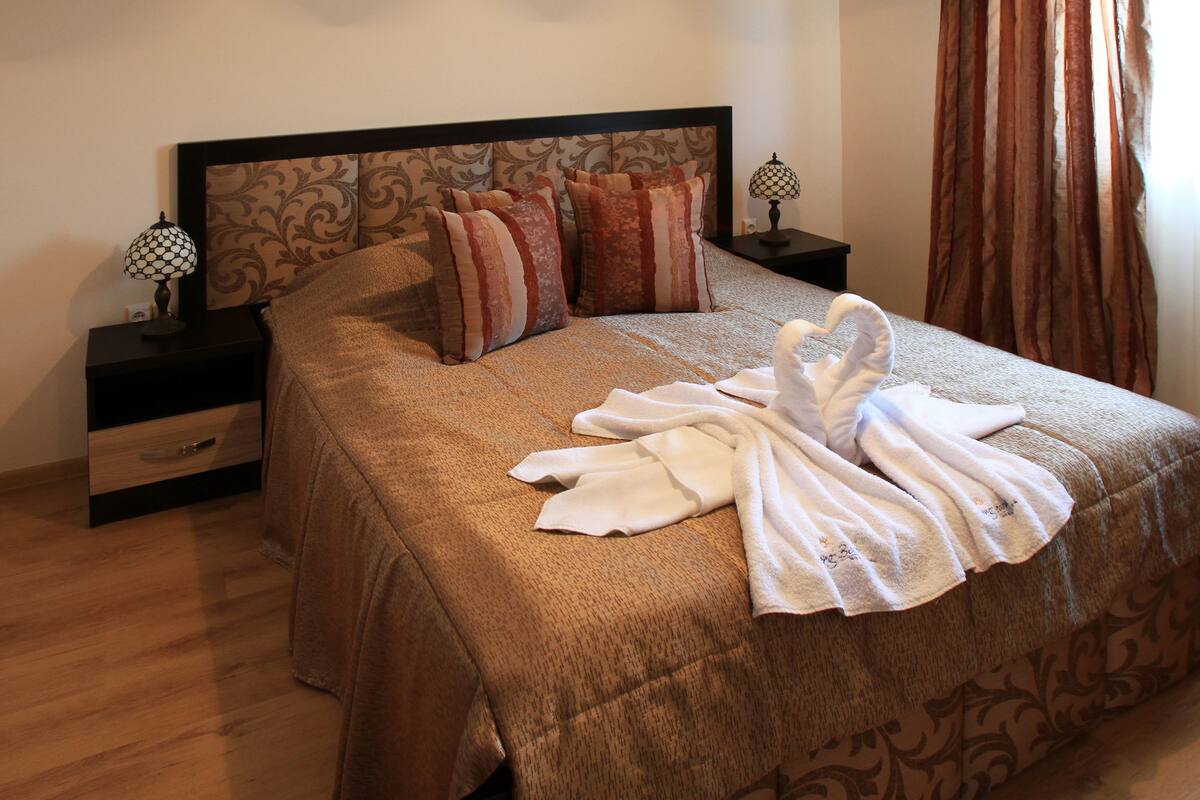 A neatly made bed with a brown bedspread, multiple pillows, and a pair of white towels. There are two bedside tables with lamps, one on each side of the bed.