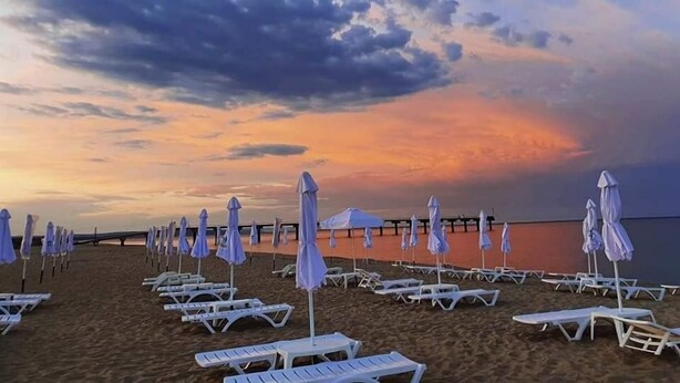 A beach with rows of white lounge chairs and blue umbrellas, under a cloudy sky. - Long Beach Resort & Spa (Dolni Chiflik)