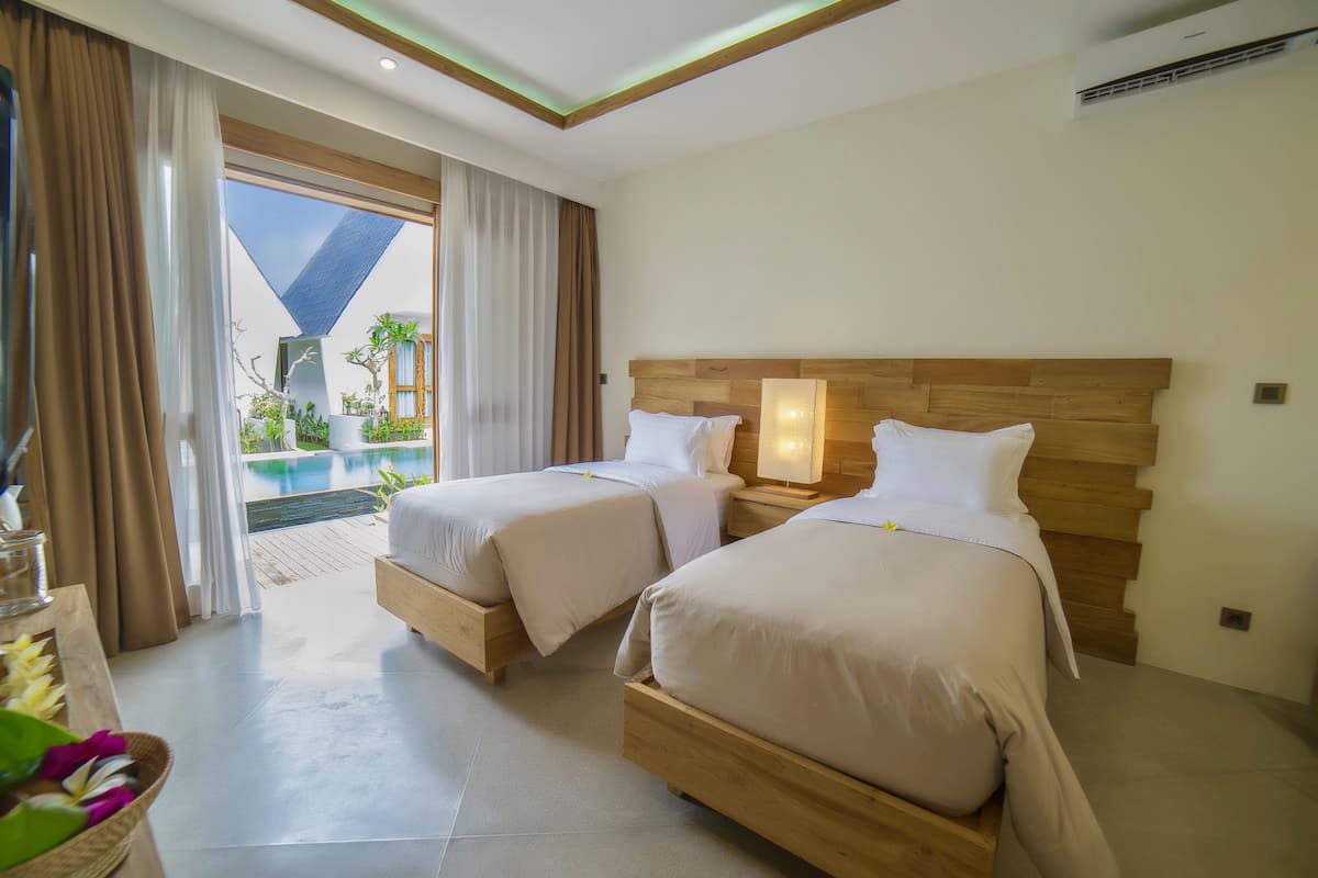 A hotel room with two beds, a wooden headboard, a nightstand with a lamp, and a view of an outdoor pool area.