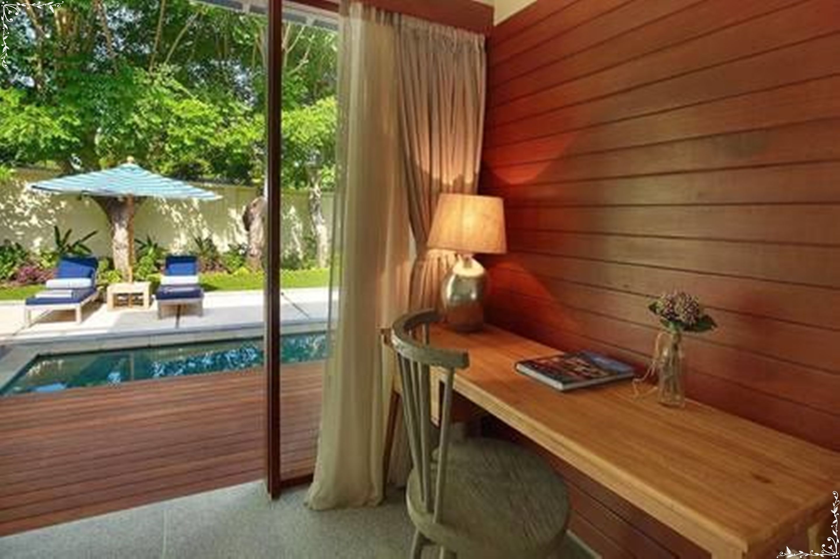 A room with a wooden desk, a chair, a lamp, and a small table with a book and a vase. There is a window with curtains, a view of a pool and outdoor seating.