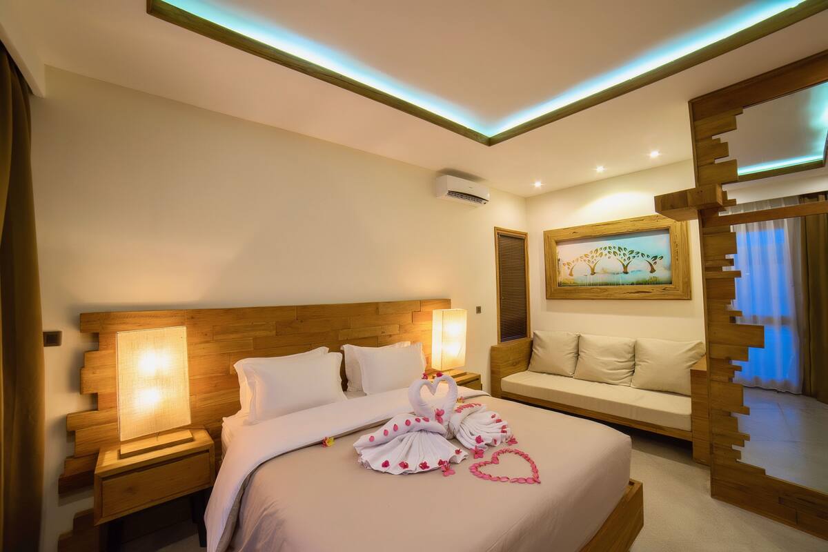 A modern hotel room with a wooden headboard, a bed with white linens, a bedside table with a lamp, a wooden bench, and a painting on the wall.