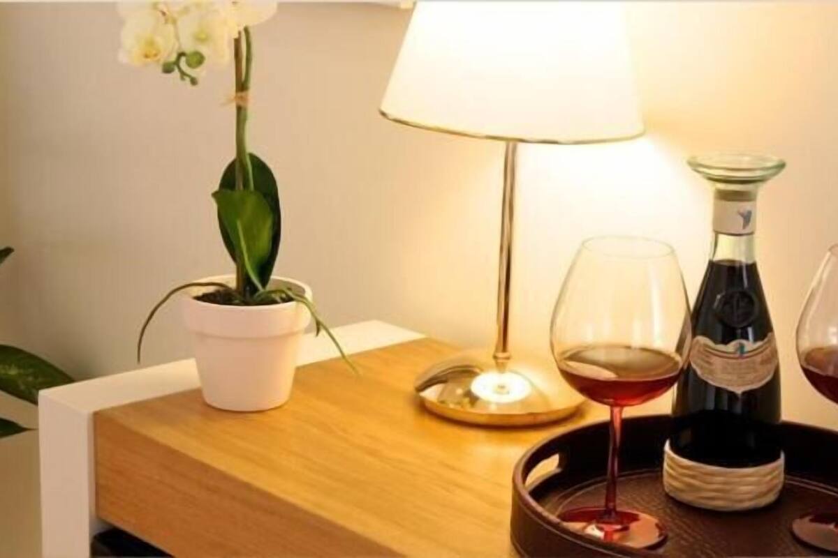 Une table en bois ornée d’une orchidée en pot, d’une lampe et de deux verres à vin, avec une bouteille de vin à proximité.