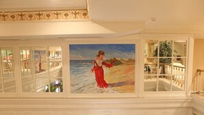 A grand hallway with a large painting of a woman in a red dress by the sea, ornate white railings, and a view of a dining area. - Hotel Gelibolu (Gallipoli)
