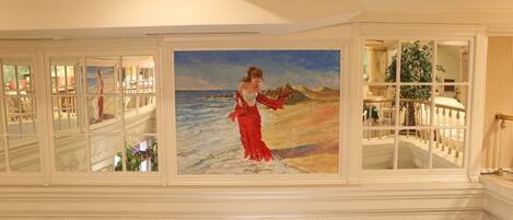 Un gran pasillo con un cuadro de una mujer en vestido rojo junto al mar, barandillas blancas ornamentadas y vistas a un comedor.