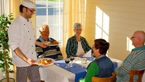 A dining room with a table set for four people, a waiter serving food, and a potted plant in the corner. - Olderfjord Turistsenter (Porsanger)