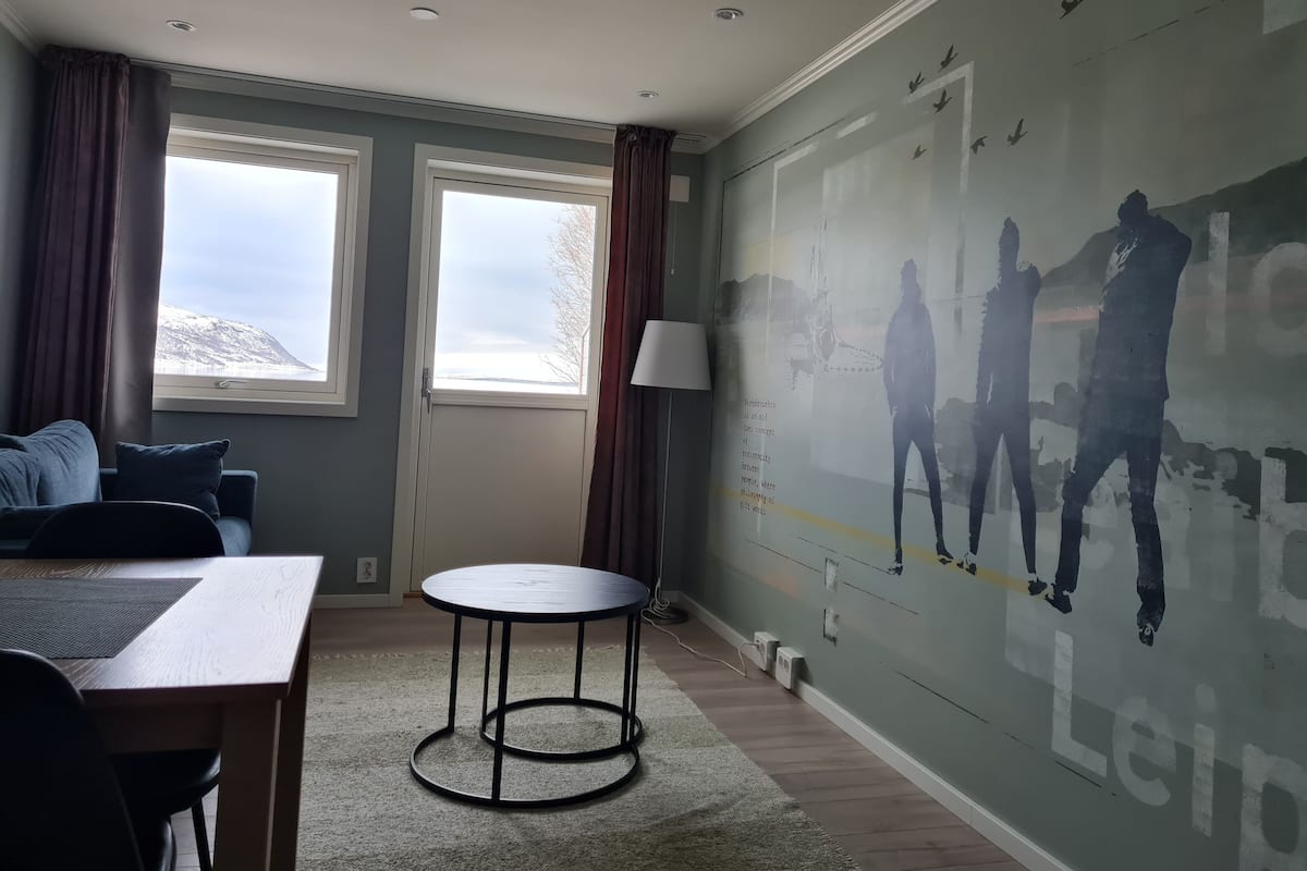 A room with a large window, a round table, a sofa, and a wall mural.