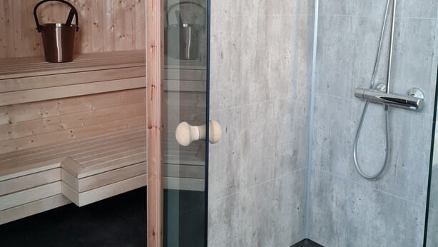 A sauna with a wooden bench and a metal bucket, a glass door leading to a shower area with a handheld showerhead and a wall-mounted faucet. - Olderfjord Turistsenter (Porsanger)