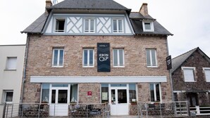 A two-story hotel with a stone facade, a sign reading "Le Bon Camp," and outdoor seating with tables and chairs.