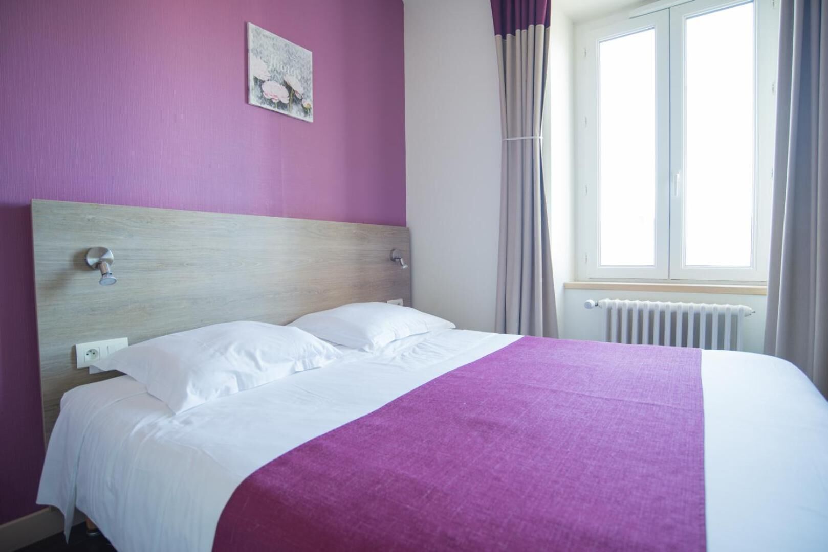Een bed met witte lakens en een paarse deken, een houten hoofdbord, een wandcontactdoos en een raam met gordijnen.