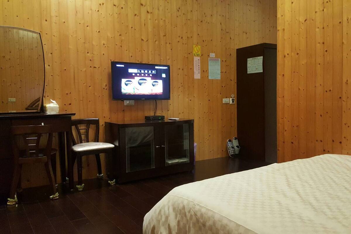 Een hotelkamer met een bed, een aan de muur bevestigde televisie, een bureau met stoel en een klein kastje.