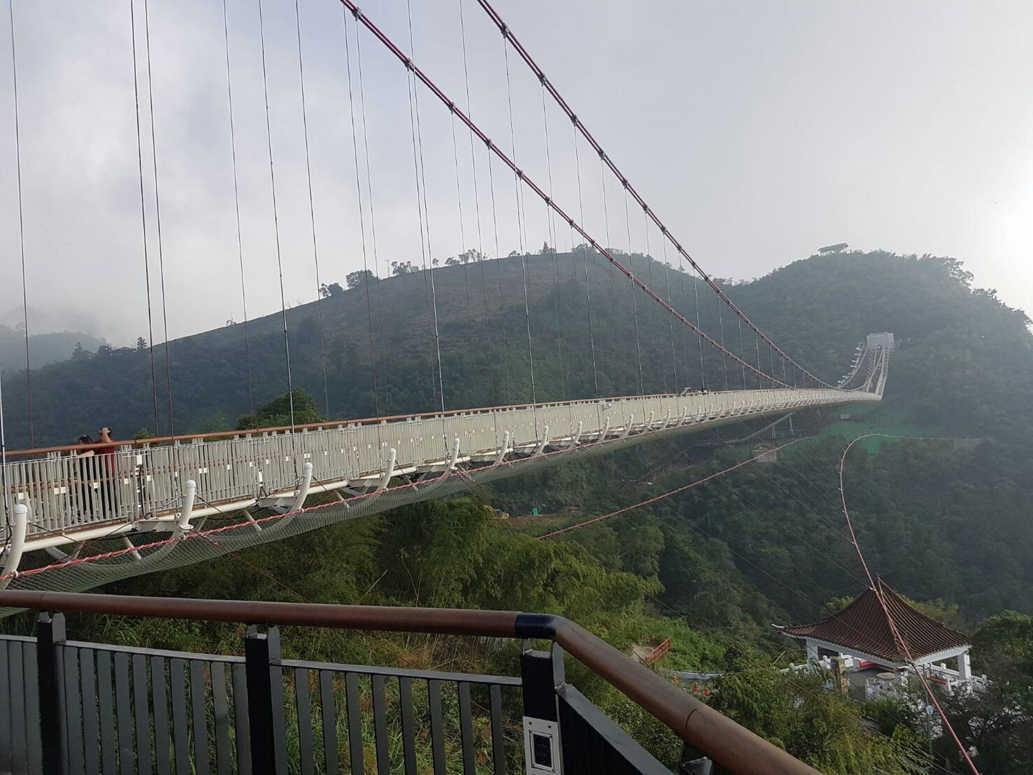 A suspension bridge with a pedestrian walkway, surrounded by lush greenery and hills.