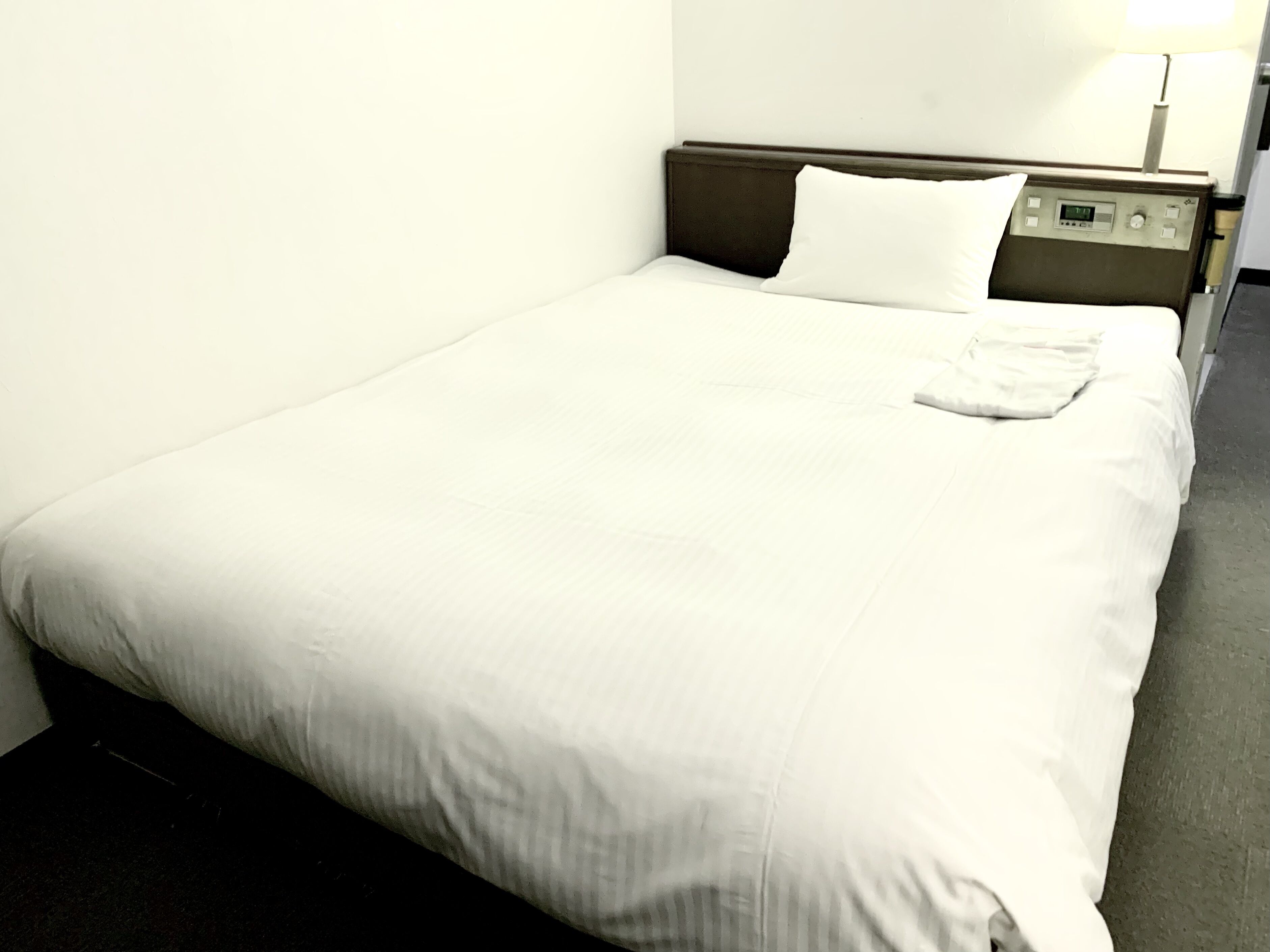 Habitación de hotel con una cama individual, cabecera con panel de control y mesita de noche con lámpara.