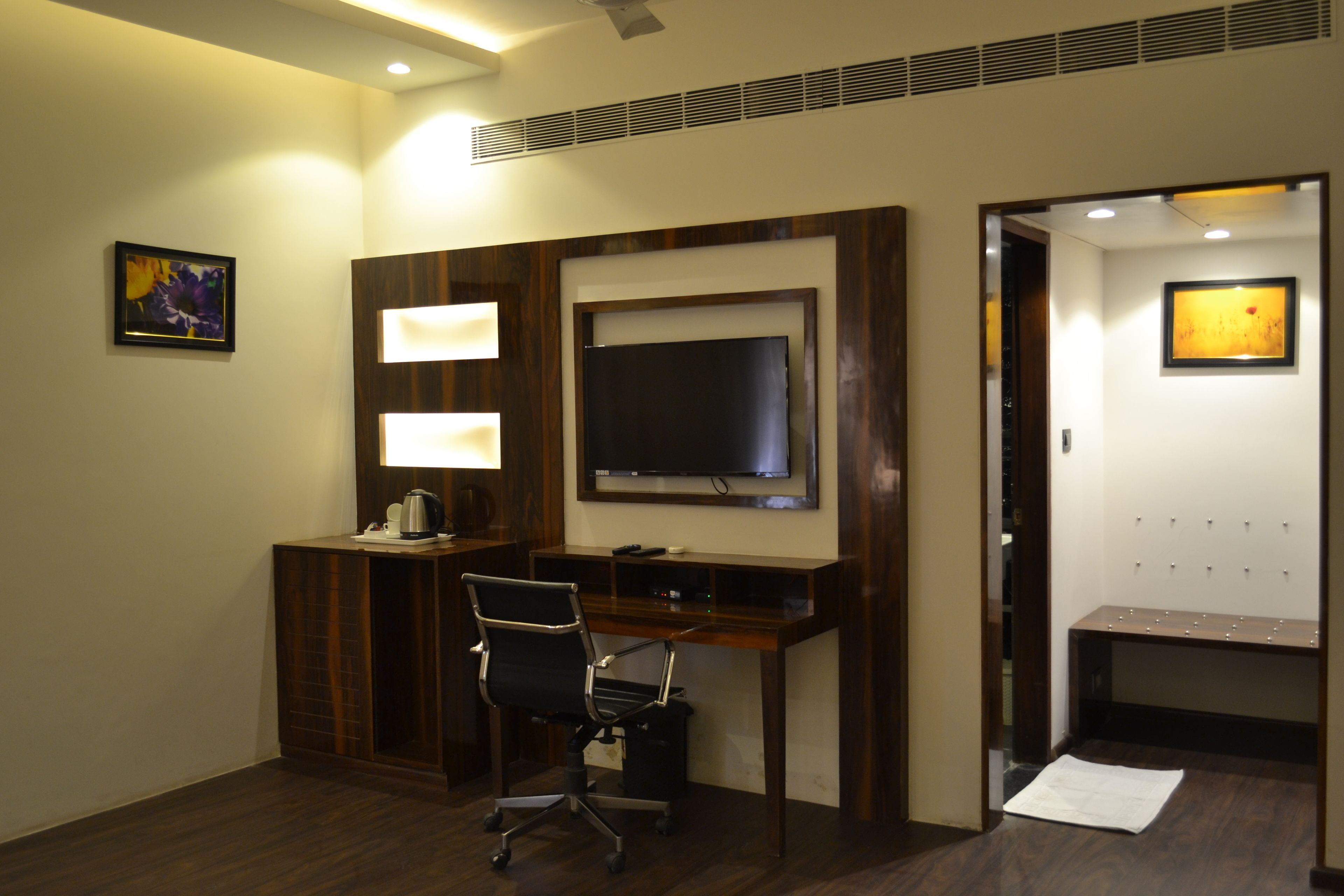 A hotel room with a wooden desk, a chair, a flat-screen TV, a coffee maker, and a coat rack.
