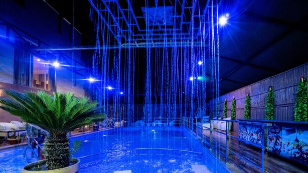 An indoor swimming pool with a water feature, surrounded by a wooden deck and potted plants. - 5 Flowers Ananta Elite, Kota (Kota)