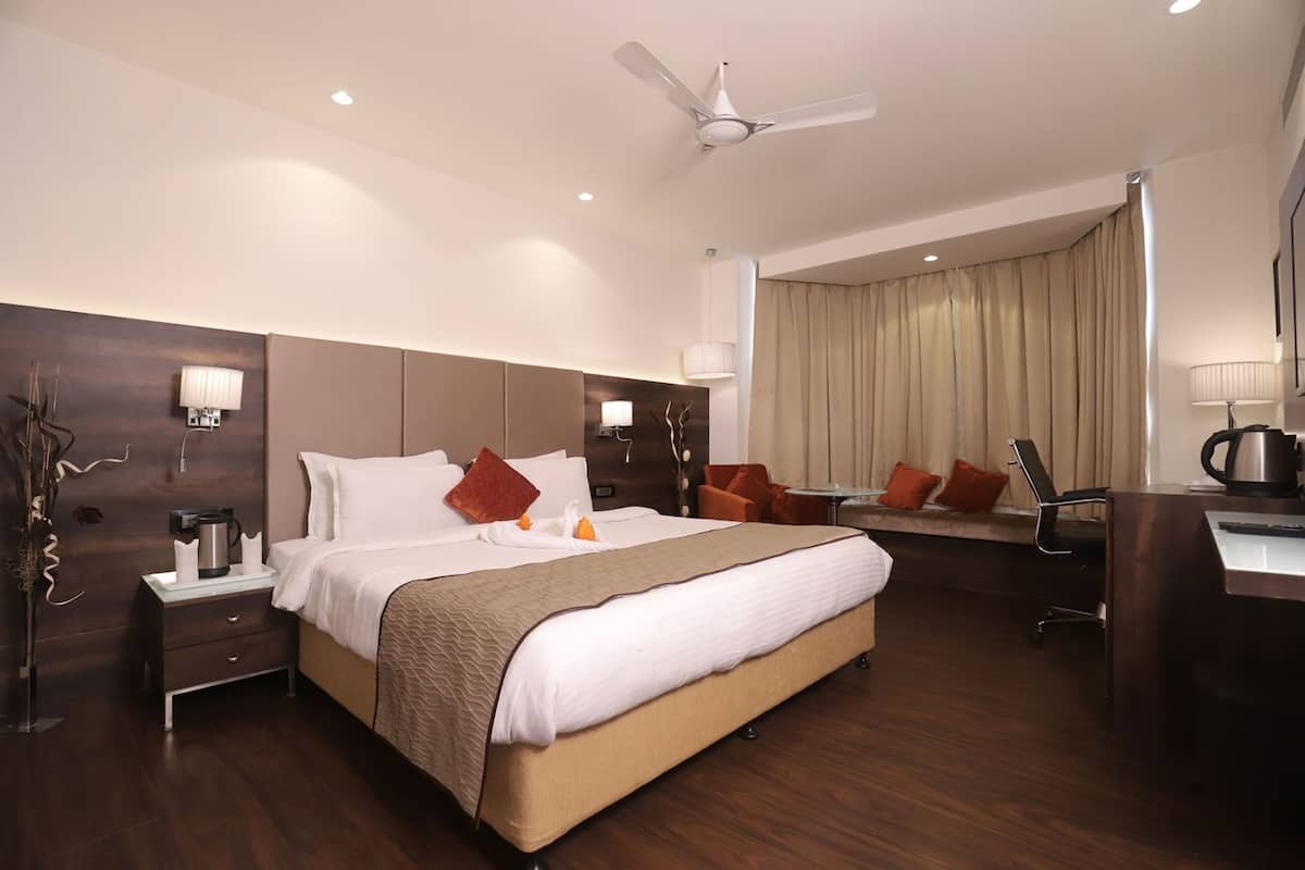 A hotel room with a bed, a desk, a chair, a coffee maker, a lamp, a ceiling fan, and a window with curtains.
