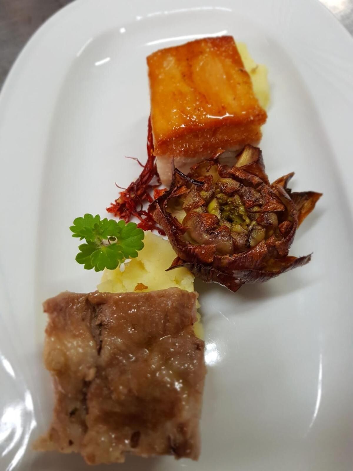 Un plato blanco con tres preparaciones diferentes: un trozo de carne glaseada, un vegetal asado y una porción pequeña de puré de papas.