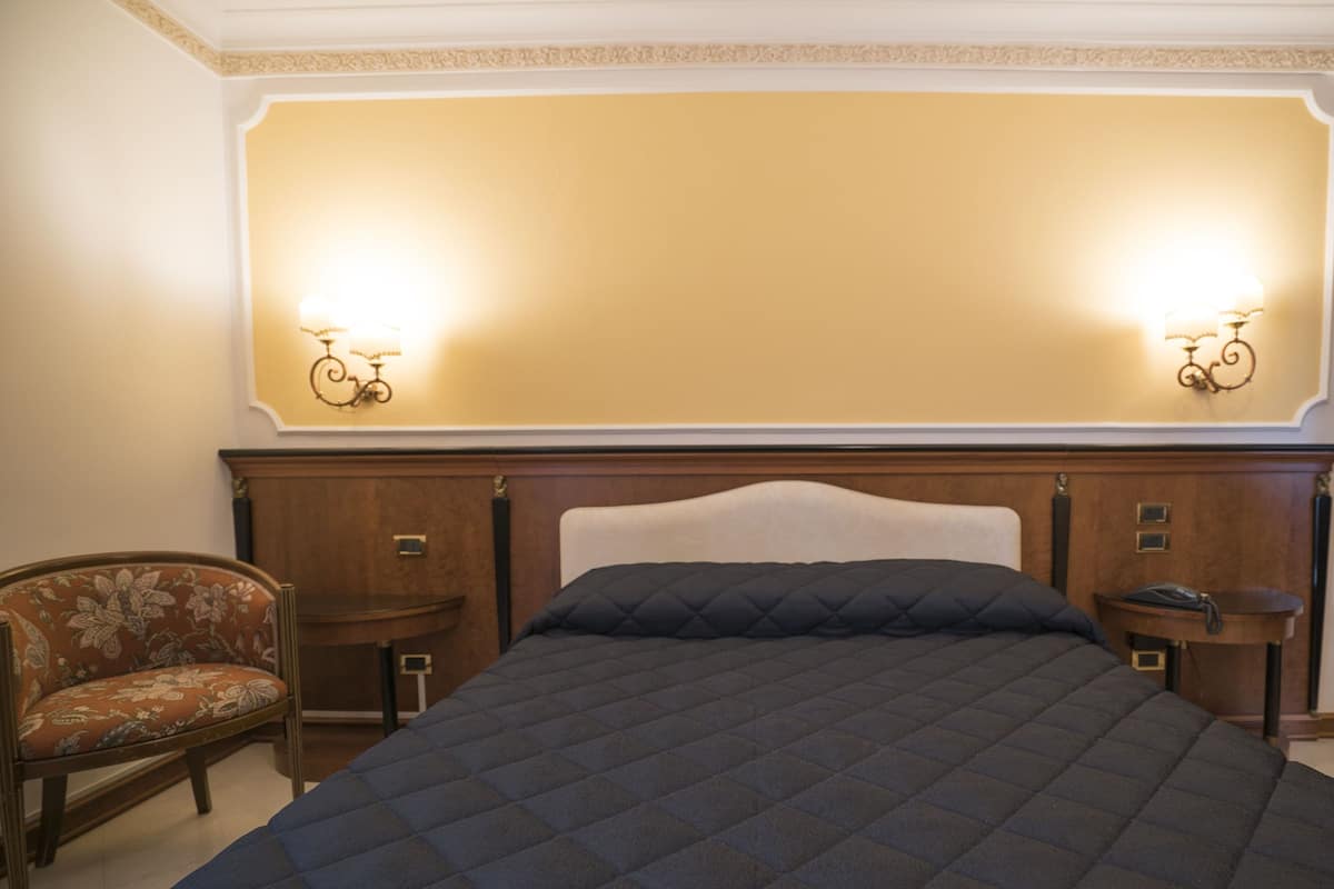 A hotel room with a large bed, two bedside tables, a chair, and wall sconces.