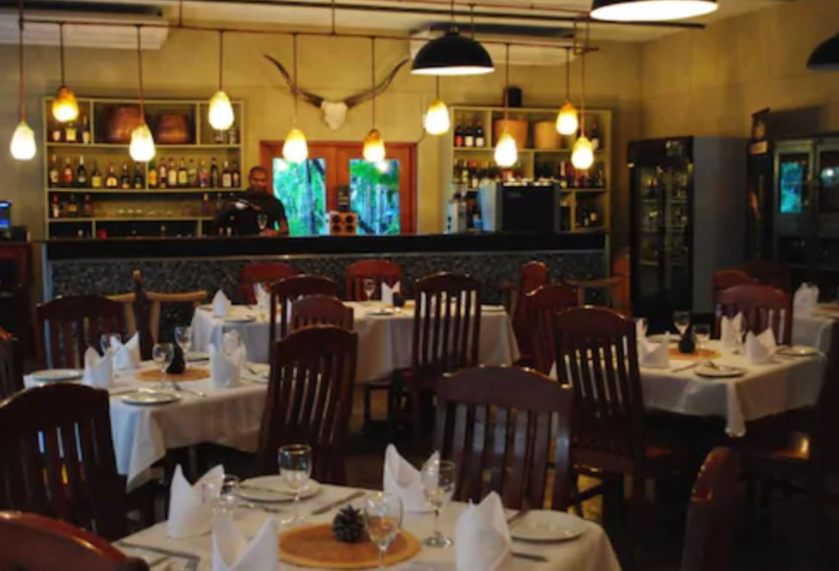 A restaurant with tables set for dining, a bar area with bottles, and a television screen.