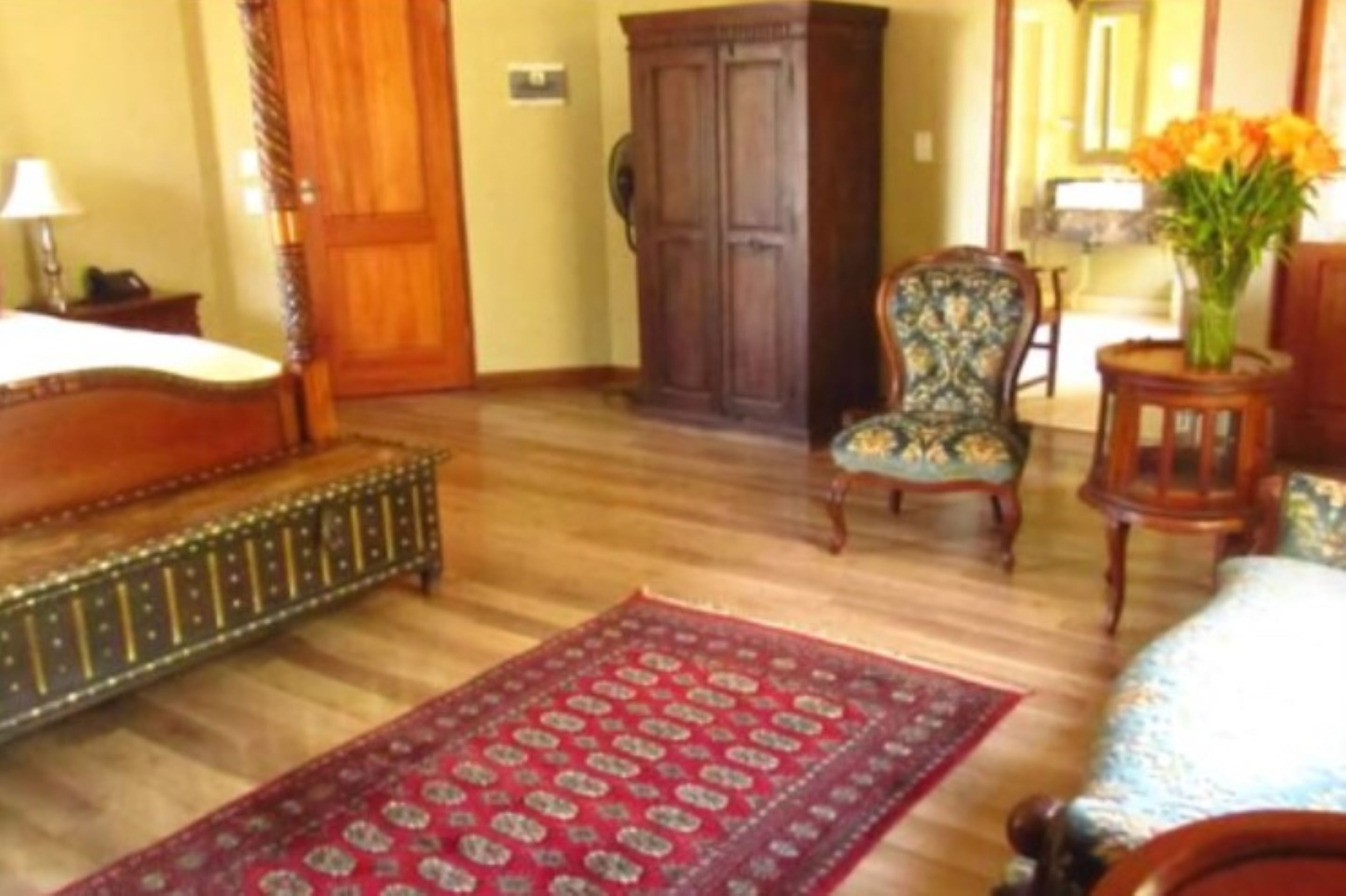 A room with a wooden floor, a bed with a metal frame, a wooden wardrobe, a patterned armchair, a small table with a vase of flowers, and a red patterned rug.