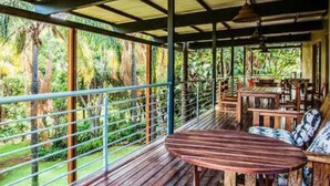 An outdoor wooden deck with metal railings, a table, and chairs, surrounded by lush greenery.