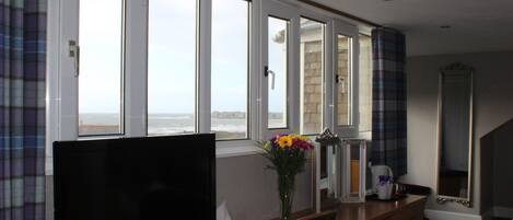 A hotel room with a large window, a television, a desk with a vase of flowers, and a mirror.