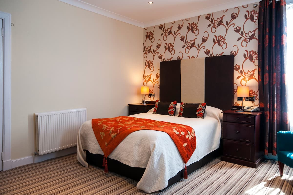A hotel room with a bed, two bedside tables with lamps, a radiator, a patterned wall, and a window with curtains.